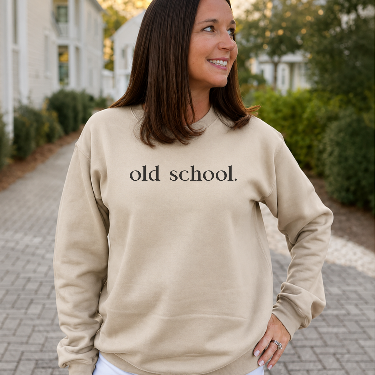 A woman wearing a beige crewneck sweatshirt with the text 'old school.' printed in black across the chest, standing outdoors on a paved walkway with houses and greenery in the background.