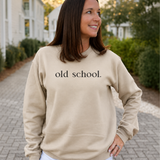 A woman wearing a beige crewneck sweatshirt with the text 'old school.' printed in black across the chest, standing outdoors on a paved walkway with houses and greenery in the background.