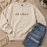 A cream-colored crewneck sweatshirt with the text 'old school.' printed in black serif font on the chest, styled flat-lay with folded blue jeans on a woven rug.