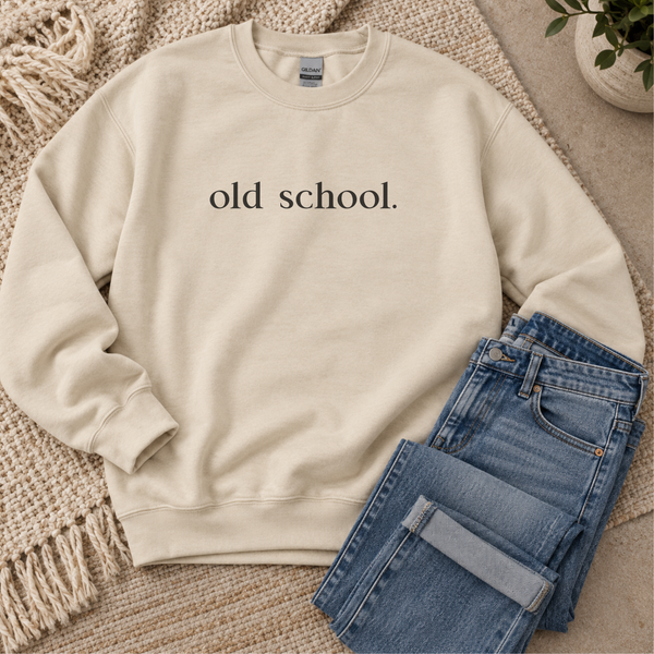 A cream-colored crewneck sweatshirt with the text 'old school.' printed in black serif font on the chest, styled flat-lay with folded blue jeans on a woven rug.