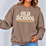 A woman wearing a taupe brown crewneck sweatshirt with a retro graphic that reads 'OLD SCHOOL' in cream letters above three horizontal stripes in cream, orange, yellow, and green. She is wearing blue jeans and a delicate gold necklace against a light gray studio background.