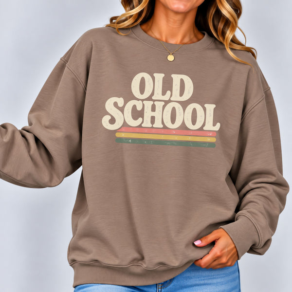 A woman wearing a taupe brown crewneck sweatshirt with a retro graphic that reads 'OLD SCHOOL' in cream letters above three horizontal stripes in cream, orange, yellow, and green. She is wearing blue jeans and a delicate gold necklace against a light gray studio background.