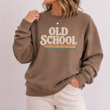 A woman wearing a brown crewneck sweatshirt with a retro-style 'OLD SCHOOL' graphic in cream and horizontal stripes, paired with off-white pants, against a light gray studio background.