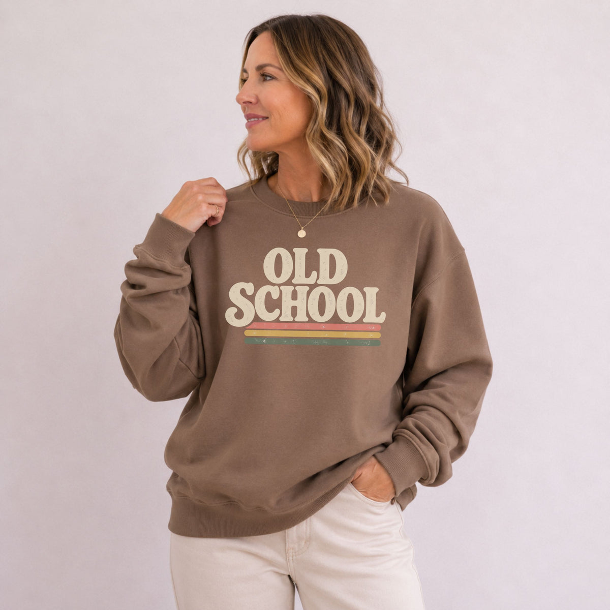 A woman wearing a brown crewneck sweatshirt with a retro-style graphic that reads 'OLD SCHOOL' in beige letters with a yellow and green stripe detail, paired with off-white pants, set against a plain light background.