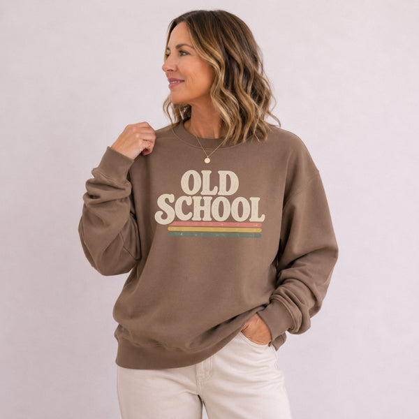 A woman wearing a brown crewneck sweatshirt with a retro-style graphic that reads 'OLD SCHOOL' in beige letters with a yellow and green stripe detail, paired with off-white pants, set against a plain light background.