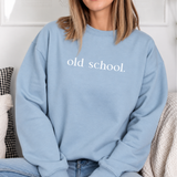 A woman wearing a light blue crewneck sweatshirt with the words 'old school.' printed in white across the chest, sitting in a cozy indoor setting.