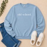 A light blue crewneck sweatshirt with the words 'old school.' printed in white across the chest, styled in a flat lay with blue jeans, white sneakers, and a sprig of greenery on a beige background.