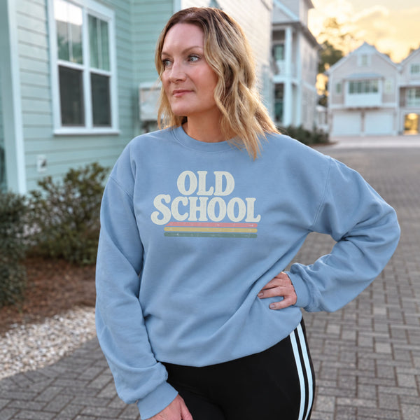 A woman wearing a light blue crewneck sweatshirt with a retro 'OLD SCHOOL' graphic, standing outdoors on a paved driveway in front of houses.