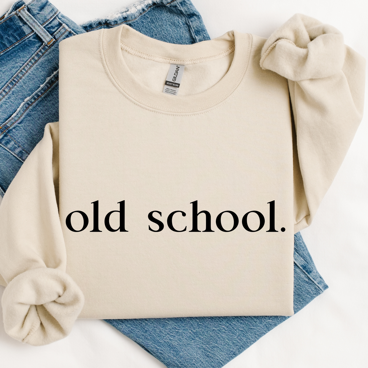 Beige sweatshirt with 'old school.' text  laying on a pair of folded jeans on a white background