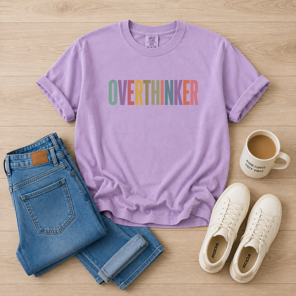 A styled flat-lay featuring a lavender purple t-shirt with the word 'OVERTHINKER' printed across the chest in multicolored letters. The t-shirt is arranged on a light wood background alongside folded blue jeans, a white coffee mug, and a pair of white sneakers.