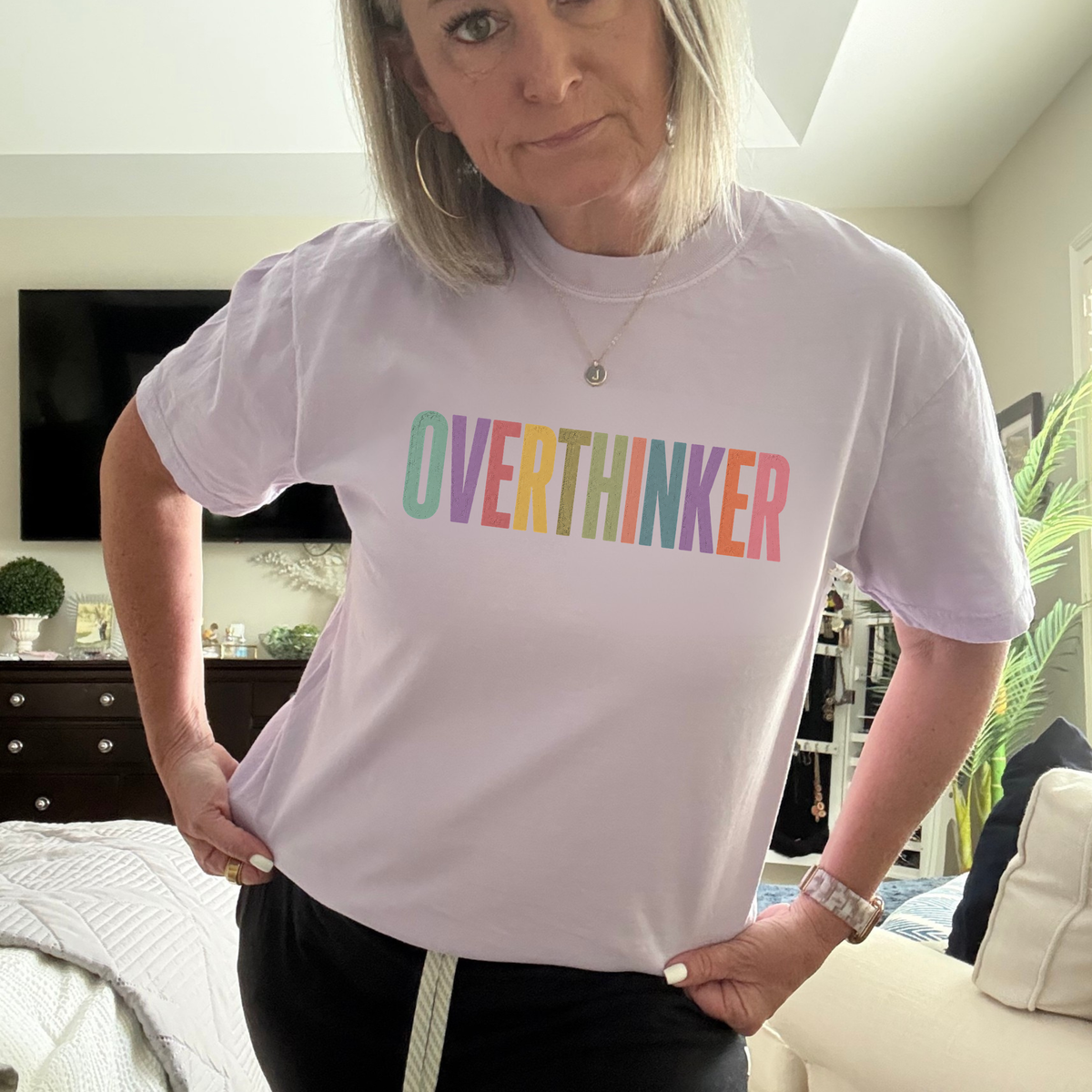 A woman wearing a light lavender crewneck t-shirt with the word 'OVERTHINKER' printed across the chest in colorful pastel letters, standing in a bedroom setting.