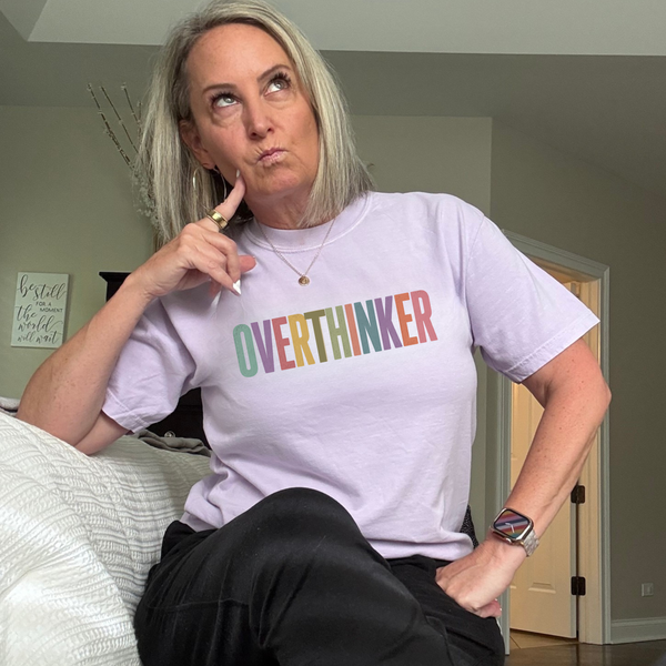 A woman sitting on a bed wearing a light lavender t-shirt with the word 'OVERTHINKER' printed across the chest in a rainbow gradient. She is posing with her hand on her chin, wearing black pants and a smartwatch.
