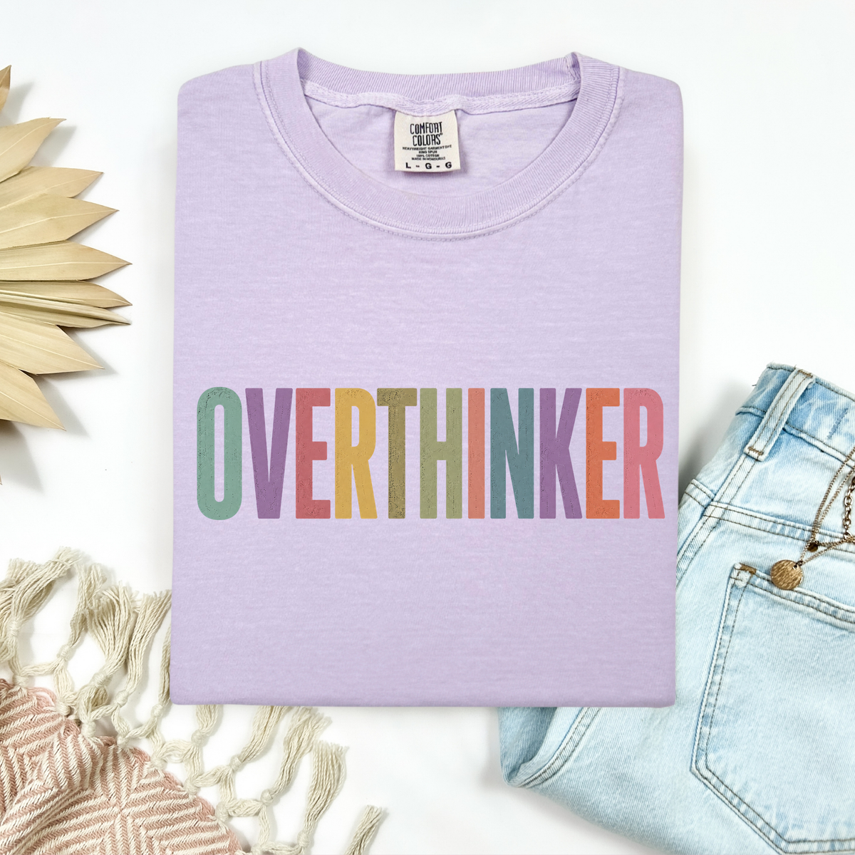 A folded light purple t-shirt with the word 'OVERTHINKER' printed across the chest in a colorful rainbow gradient. The shirt is styled in a flat-lay arrangement with light blue jeans, a beige woven textile, and a wooden sun decoration on a white background.
