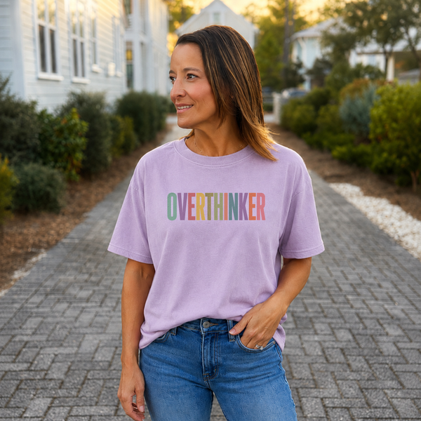A woman wearing a light purple t-shirt with the word 'OVERTHINKER' printed across the chest in pastel rainbow colors, standing on a paved path in front of houses.