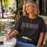 A woman sitting at an outdoor cafe table wearing a dark charcoal t-shirt with the word 'OVERTHINKER' printed across the chest in a colorful rainbow gradient. She is holding a glass of white wine, with a blurred cafe background.