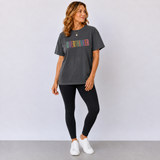 A woman modeling a dark gray oversized graphic t-shirt with the word 'OVERTHINKER' printed across the chest in a multicolor gradient. She is wearing black leggings and white sneakers against a light gray studio background.
