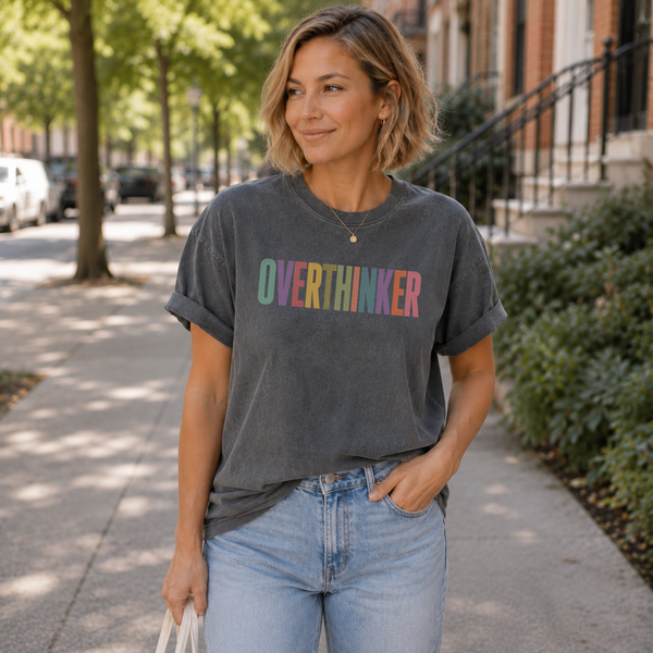 A woman standing on a city sidewalk wearing a faded dark gray t-shirt with the word 'OVERTHINKER' printed across the chest in pastel rainbow colors, paired with light blue jeans.
