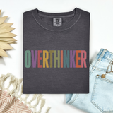A folded dark gray t-shirt with the word 'OVERTHINKER' printed across the chest in colorful retro letters, styled in a flat lay with light blue jeans, a woven textile, and a wooden sun decoration on a white background.