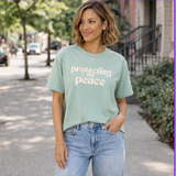 Woman wearing a light green t-shirt with 'protecting my peace' text, standing on a sidewalk.