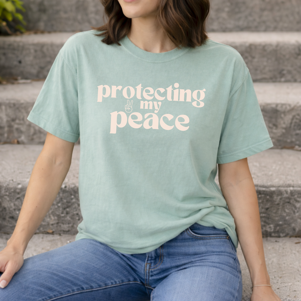 A woman wearing a light green t-shirt with 'protecting my peace' text, sitting on steps.