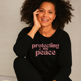 Woman wearing a black sweatshirt with 'protecting my peace' text, sitting against a neutral background.