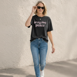 A Woman wearing a black t-shirt with 'protecting my peace' text and blue jeans against a light background
