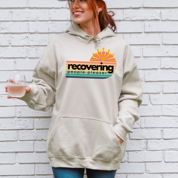 Person wearing a beige hoodie with a colorful graphic and text, standing against a white brick wall.