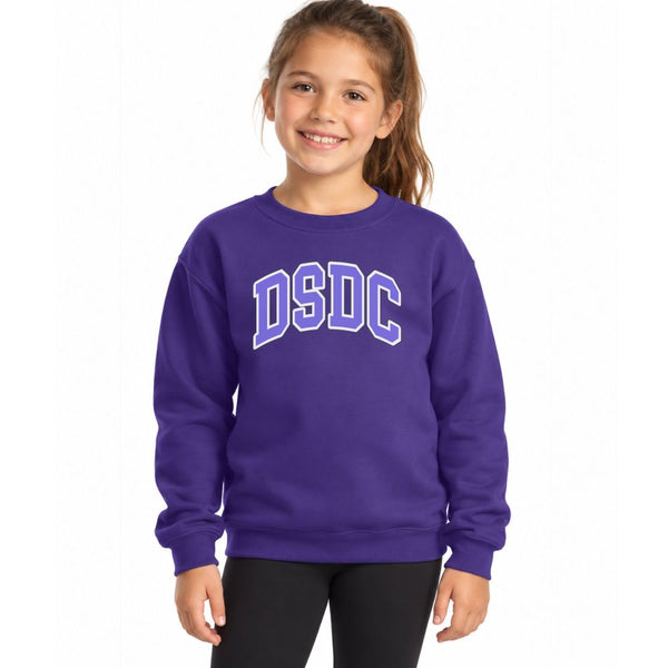 A young girl with her hair in a ponytail smiling while wearing a purple crewneck sweatshirt with 'DSDC' printed in white and purple block letters across the chest, paired with black pants, against a plain white background.