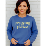 Woman wearing a blue sweatshirt with 'protecting my peace' in yellow text against a white brick wall.
