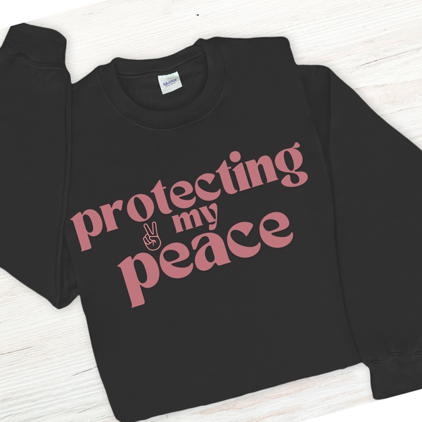 Black sweatshirt with pink text 'protecting my peace' on a white wooden surface