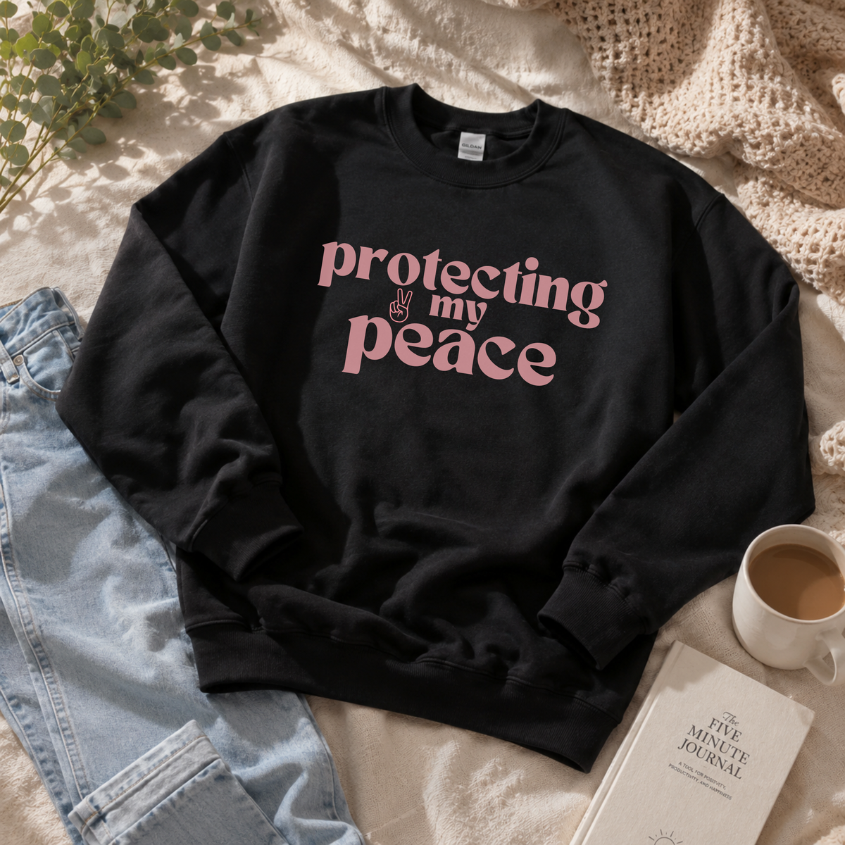 A black crewneck sweatshirt with the phrase 'protecting my peace' printed in pink retro-style lettering across the chest, styled in a cozy flat-lay arrangement with light blue jeans, a cup of coffee, a book, and a knit blanket.