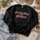 A black crewneck sweatshirt with the phrase 'protecting my peace' printed in pink retro-style lettering across the chest, styled in a cozy flat-lay arrangement with light blue jeans, a cup of coffee, a book, and a knit blanket.