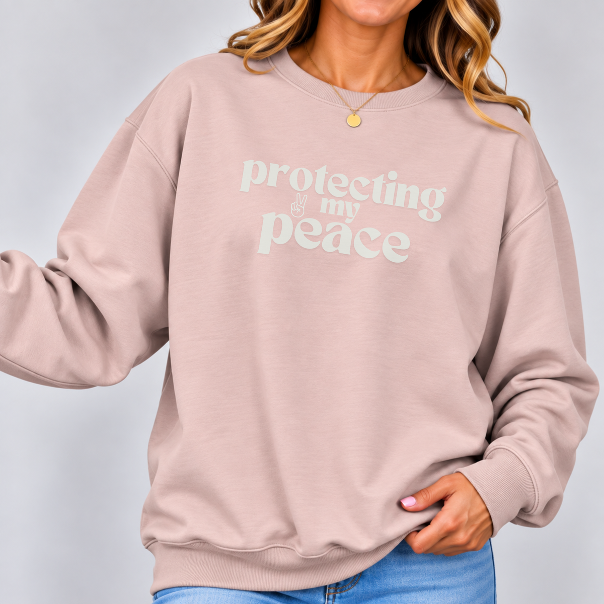 A woman wearing a dusty pink crewneck sweatshirt with the words 'protecting my peace' printed in a light mint green retro font across the chest, paired with blue jeans against a light gray studio background.