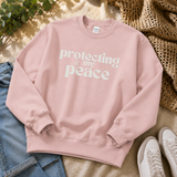 A dusty pink crewneck sweatshirt with the text 'protecting my peace' printed in white across the chest, styled in a flat lay with blue jeans, a woven blanket, and white sneakers.
