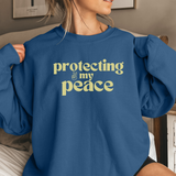 Person wearing a blue sweatshirt with 'protecting my peace' text in lemon yellow in a home setting