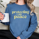 Person wearing a blue sweatshirt with 'protecting my peace' text in lemon yellow, holding a white mug.