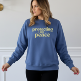 Woman wearing a blue sweatshirt with 'protecting my peace' in yellow text against a white wall.