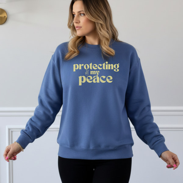 Woman wearing a blue sweatshirt with 'protecting my peace' in yellow text against a white wall.