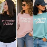 Three women modeling the same slogan sweatshirt in different colors: black with pink text, light pink with white text, and mint green with white text. Each model is wearing blue jeans and styled outdoors or in a doorway.