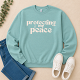 A mint green crewneck sweatshirt with the white text 'protecting my peace' and a peace sign graphic, styled in a flat lay with blue jeans, white sneakers, and a sprig of green leaves on a beige background.