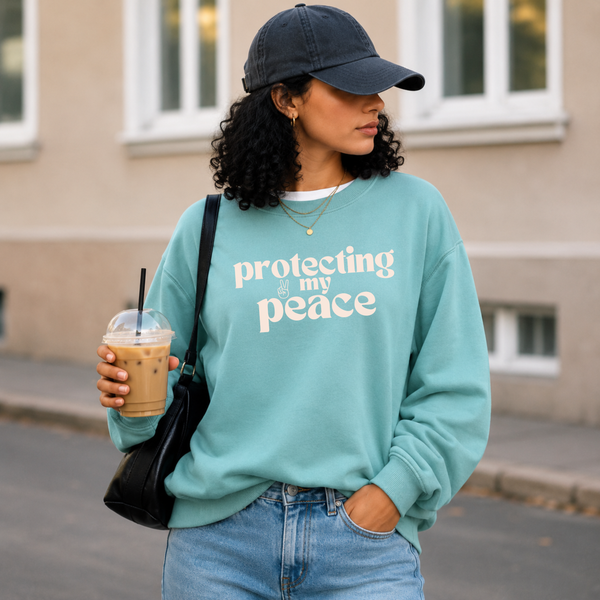 A woman wearing a light teal sweatshirt with the text 'protecting my peace' in white, paired with a dark baseball cap, blue jeans, and a black shoulder bag while holding an iced coffee outdoors.