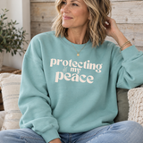 A woman wearing a sage green crewneck sweatshirt with the text 'protecting my peace' in white, sitting on a couch in a cozy room setting.