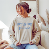 A woman wearing a beige sweatshirt with 'protecting my peace' text in blue text, sitting on a couch.