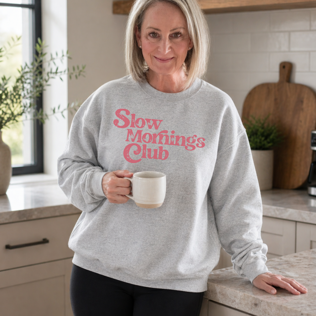 Woman wearing a gray sweatshirt with 'Slow mornings club' text, holding a mug in a kitchen.