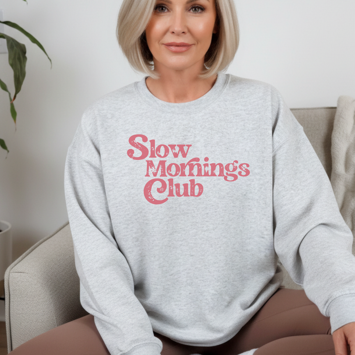 A woman wearing an ash gray sweatshirt with 'Slow Morning's Club' text in pink.