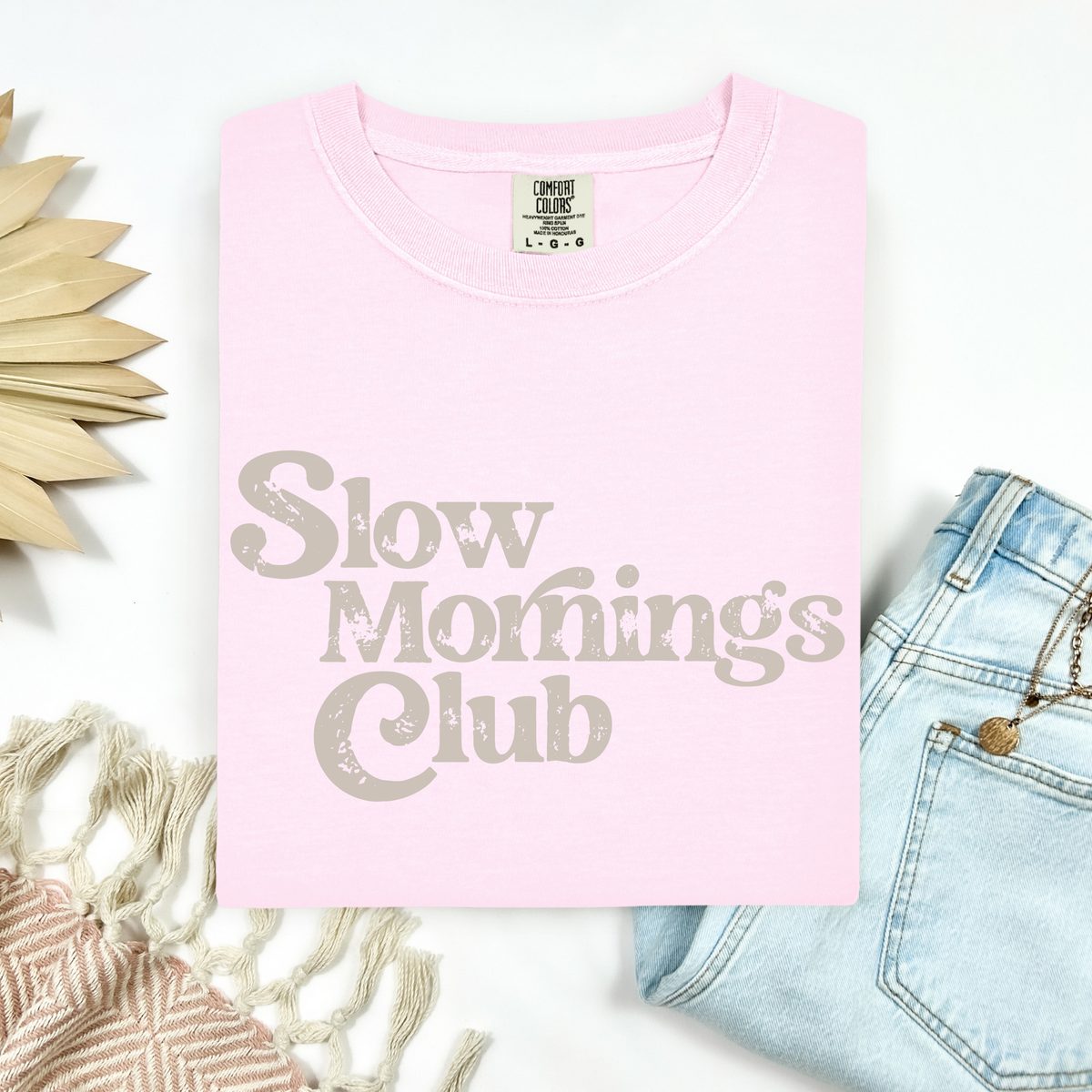 Pink t-shirt with 'Slow Mornings Club' text on a white background with accessories.