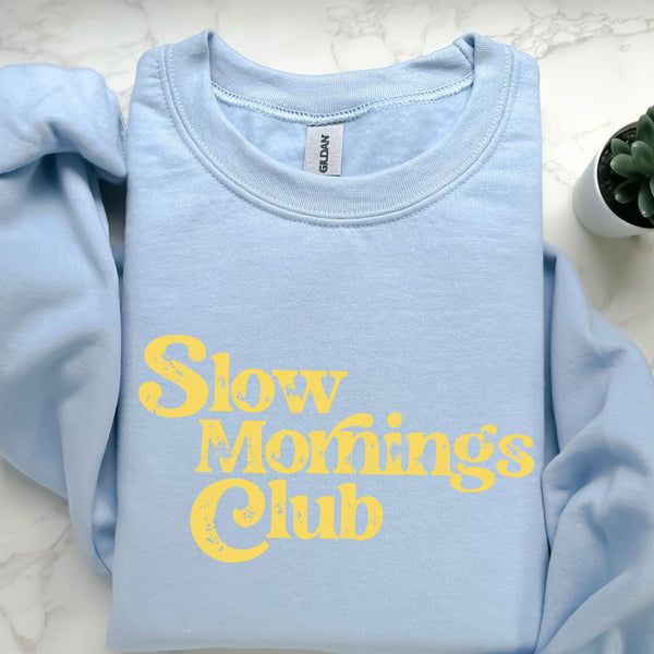 Light blue sweatshirt with yellow text 'Slow Mornings Club' on a white marble surface.
