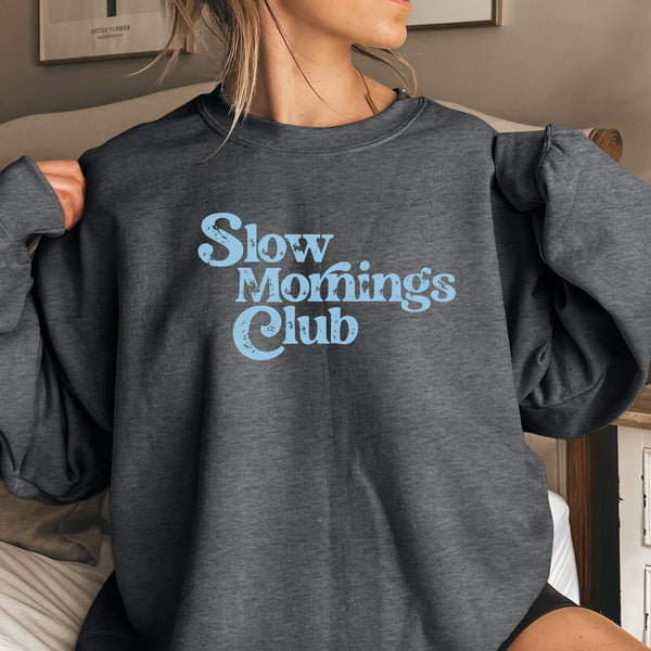 Woman  wearing a dark gray sweatshirt with 'Slow Mornings Club' text in light blue.