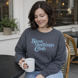 Woman wearing a 'Slow Mornings Club' sweatshirt holding a mug in a cafe setting.
