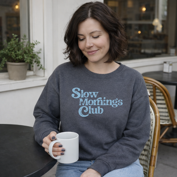 Woman wearing a 'Slow Mornings Club' sweatshirt holding a mug in a cafe setting.